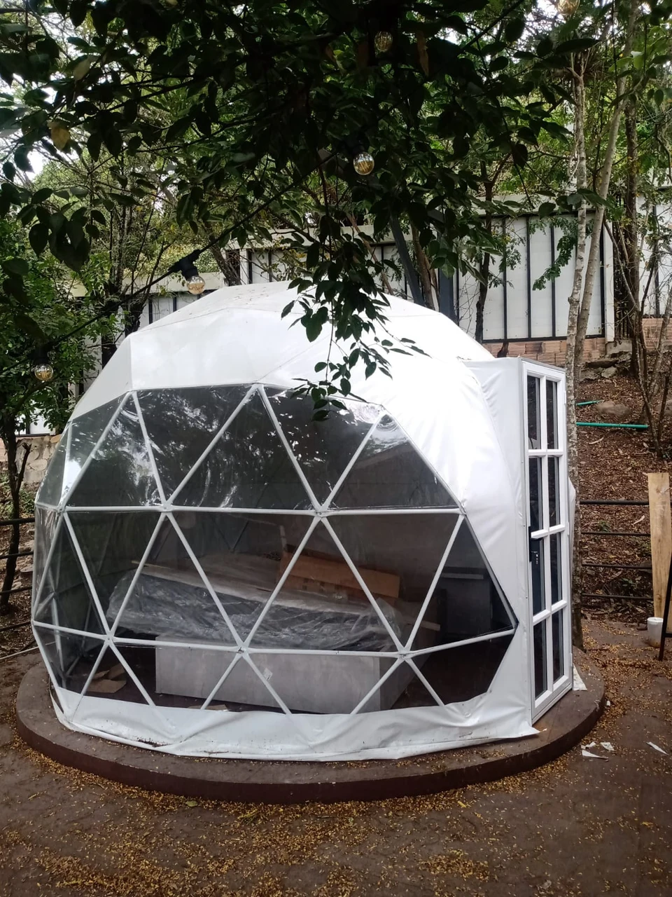 Domo geodésico de glamping terminado, con ventanal panorámico transparente en la mitad frontal, puerta de vidrio con marco blanco y lona clara sobre el resto, montado sobre una base circular de madera