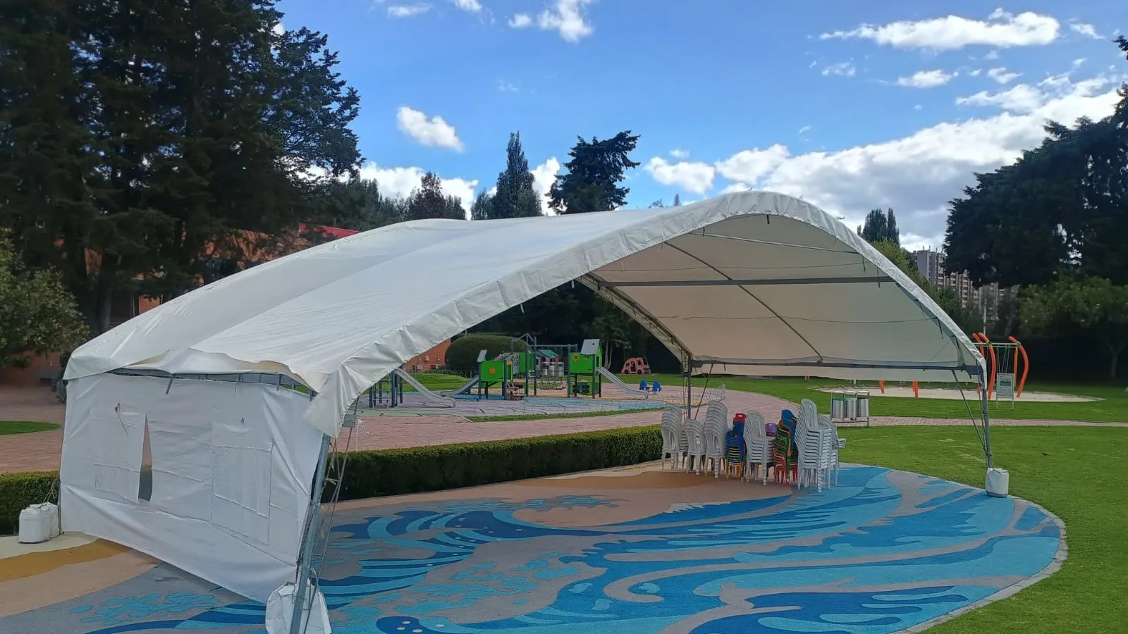 Carpa en arco de lona blanca montada sobre la plazoleta de un parque infantil, con estructura metálica curva y cerramiento lateral