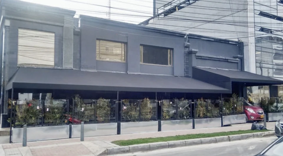 Toldo fijo de lona oscura corrido a lo largo de la terraza de un local, sobre paneles de vidrio que cierran la zona hacia la calle